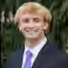 Smiling young man with blond hair wearing a dark suit, white shirt, and purple tie standing outdoors with green foliage background.