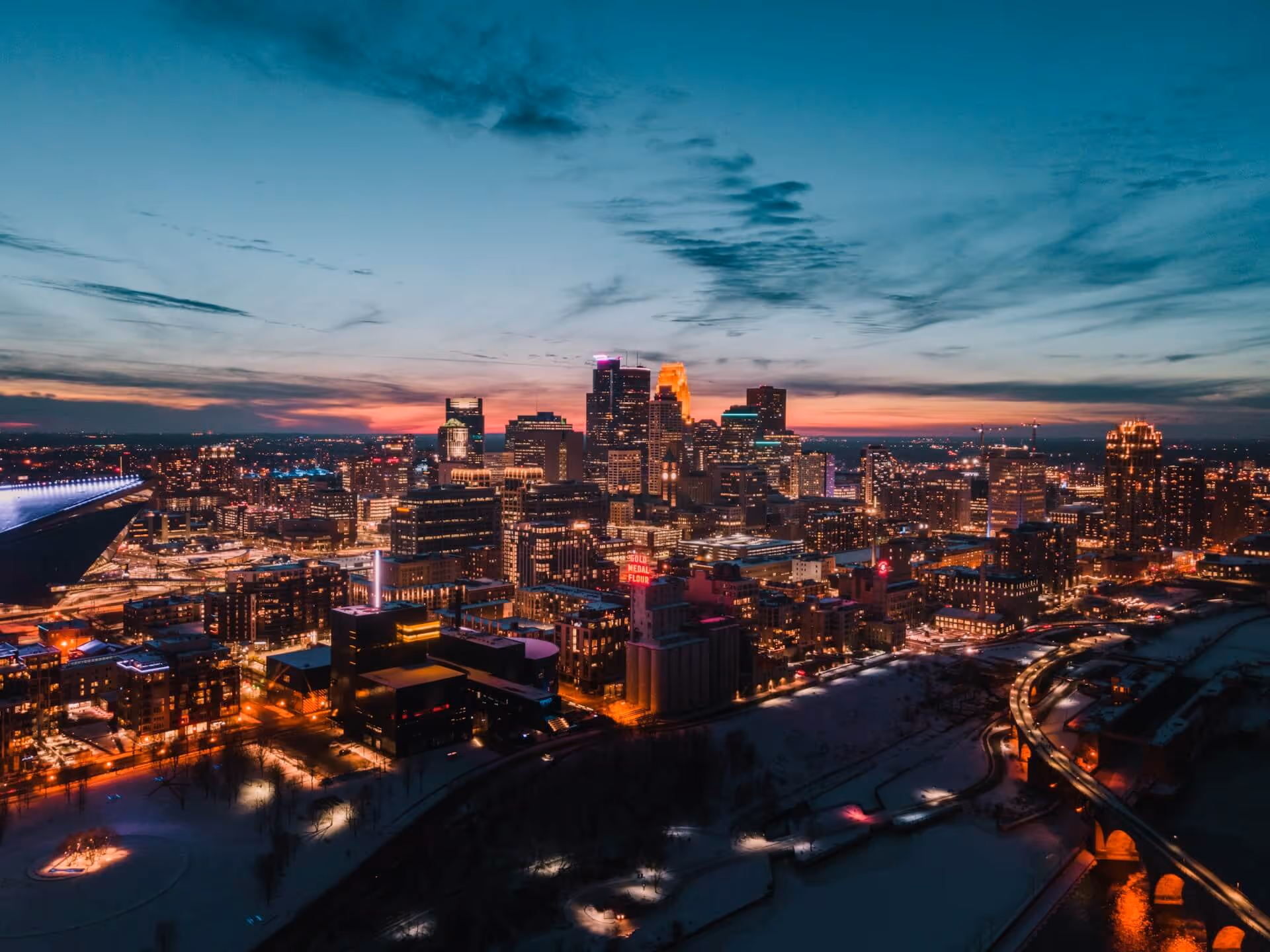 Aerial view of a city skyline at dusk with illuminated buildings and snowy streets below a blue and pink sky.