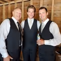 Three men dressed in black vests and white shirts standing closely inside a wooden structure, smiling at the camera.