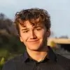 Young man with curly brown hair smiling outdoors with blurred natural background.